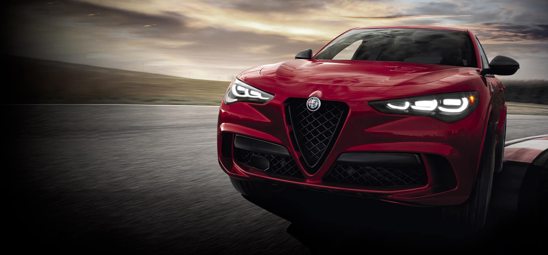 The front end of a red 2024 Alfa Romeo Stelvio Quadrifoglio being driven on a track with dramatic dark skies above.