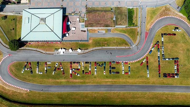 Un ángulo aéreo de un museo y una pista de carreras en Italia, con cientos de vehículos Alfa Romeo acomodados abajo para formar las palabras "Alfa Romeo 115".
