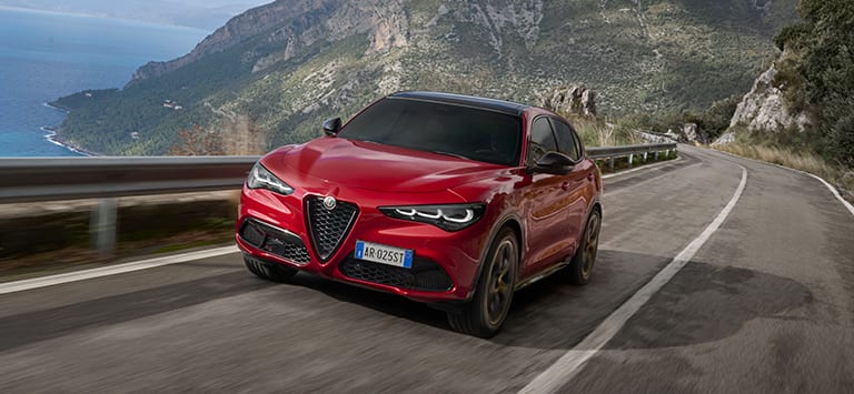 A front angle of a European version red 2025 Alfa Romeo Stelvio Intensa traveling on a highway in the mountains.
