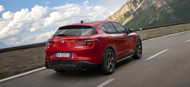 A rear angle of a European version red 2025 Alfa Romeo Stelvio Intensa traveling on a highway in the mountains.
