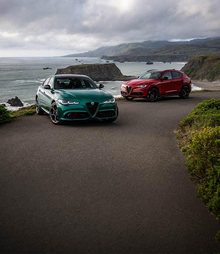 A green Alfa Romeo Giulia and a red Alfa Romeo Stelvio parked on a scenic coastal road, with rocky cliffs and the ocean in the background, during a cloudy day.