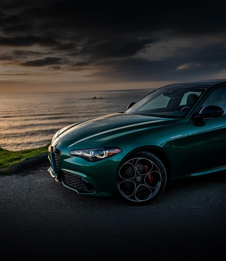 The driver-side front end of a green 2025 Alfa Romeo Giulia with Veloce Package parked on a rocky beach beside the ocean.