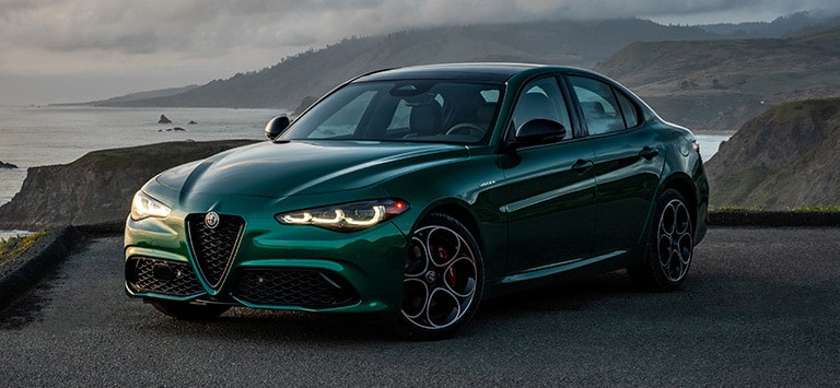 Ángulo delantero del lado del conductor de un Alfa Romeo Giulia Veloce 2026 verde estacionado en una playa rocosa.