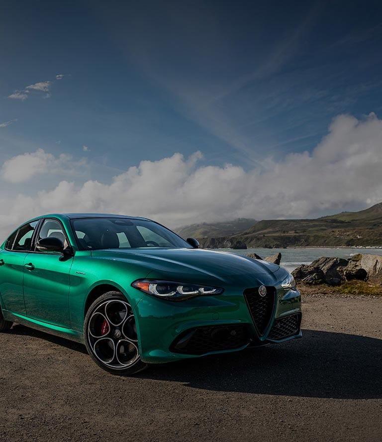 Ángulo delantero del lado del pasajero de un Alfa Romeo Giulia Veloce 2026 verde estacionado en una playa rocosa, con ominosas nubes de tormenta de fondo.