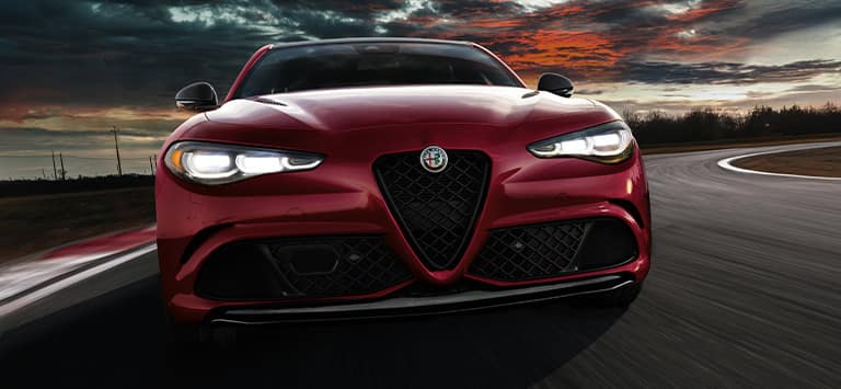 Ángulo delantero de un Alfa Romeo Giulia Veloce 2026 rojo con los faros delanteros encendidos, circulando por una pista con curvas al atardecer.