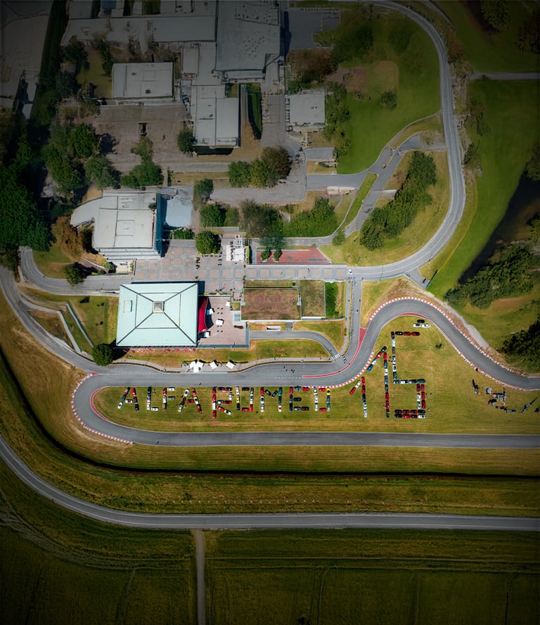 An aerial angle of a museum and racetrack in Italy with hundreds of Alfa Romeo vehicles arranged on the grounds, spelling out the words "Alfa Romeo 115".