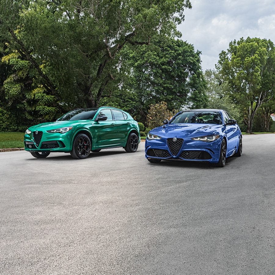 Un Alfa Romeo Stelvio Veloce 2022 azul y un Alfa Romeo Giulia Veloce 2022 blanco estacionados en la entrada de una casa de estilo contemporáneo con palmeras en el fondo.