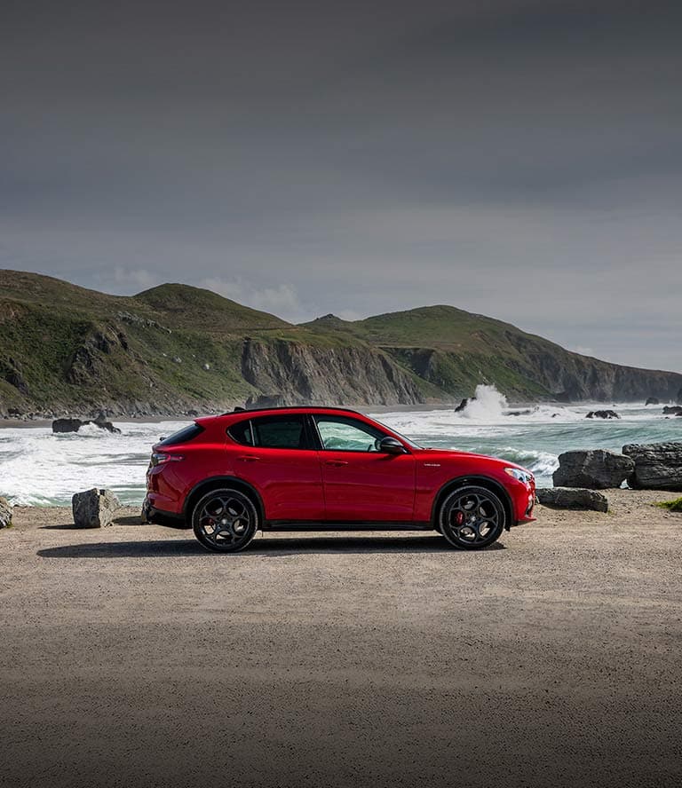 A passenger-side profile of a red 2025 Alfa Romeo Stelvio AWD with Veloce Package  parked on a rocky beach beside the ocean.