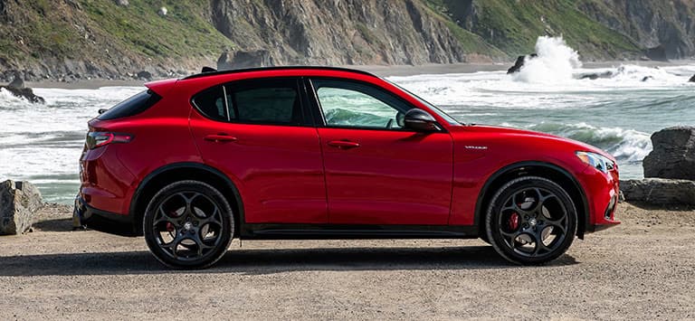 Perfil del lado del pasajero de un Alfa Romeo Stelvio Veloce 2026 rojo estacionado en una playa rocosa.