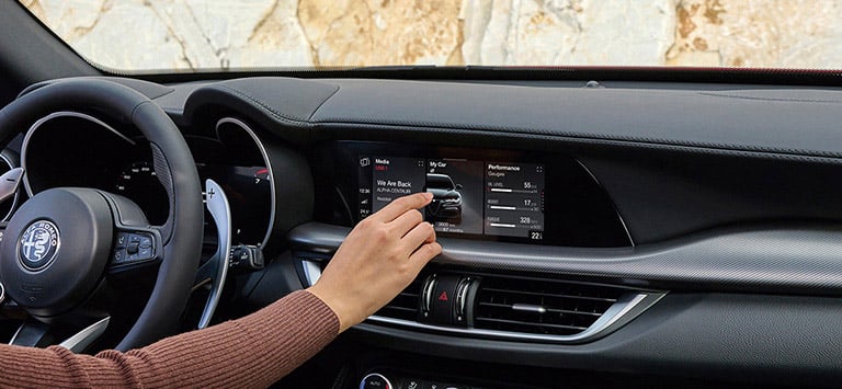 El volante, la pantalla táctil de Uconnect y las aberturas de calefacción y ventilación de un Alfa Romeo Stelvio 2026, con el conductor tocando la pantalla.