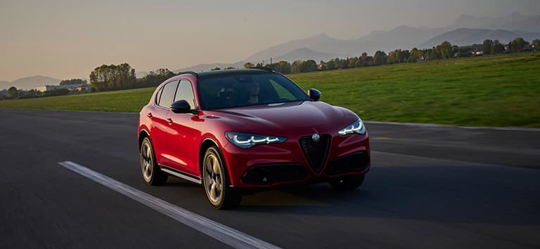 Un Alfa Romeo Stelvio Veloce 2026 rojo con los faros delanteros encendidos, circulando por una carretera al atardecer.