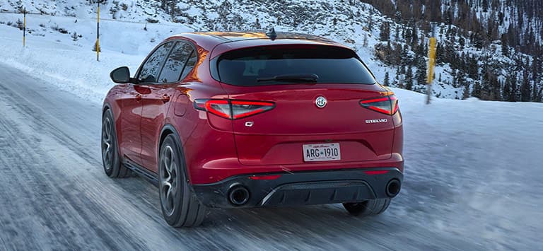 Ángulo trasero de un Alfa Romeo Stelvio Veloce 2026 rojo circulando por un camino cubierto de nieve.