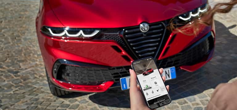 The front end of a red 2026 Alfa Romeo Tonale Sport Speciale and a woman holding a smartphone that is displaying a page from the My Alfa Romeo app.