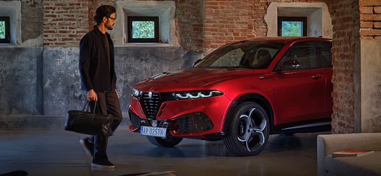 A red 2026 Alfa Romeo Tonale Sport Speciale parked with its headlamps on, parked between two brick buildings, with a man standing in front of the vehicle.