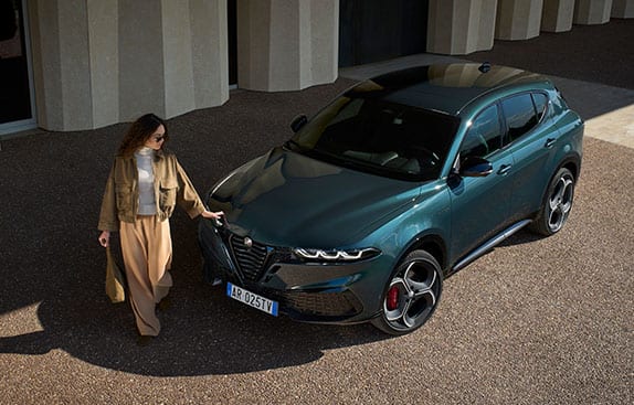 A raised angle of a green 2026 Alfa Romeo Tonale Sport Speciale parked beside a commercial building with a woman standing, touching the hood.