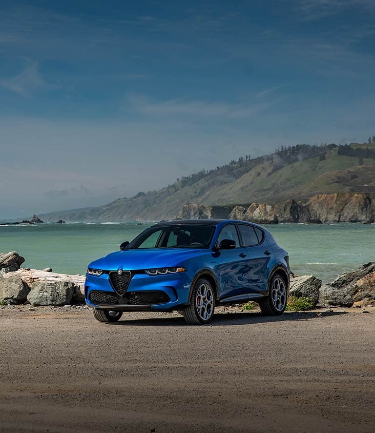 A driver-side front angle of a blue 2025 Alfa Romeo Tonale AWD with Veloce Package parked on a rocky beach beside the ocean.