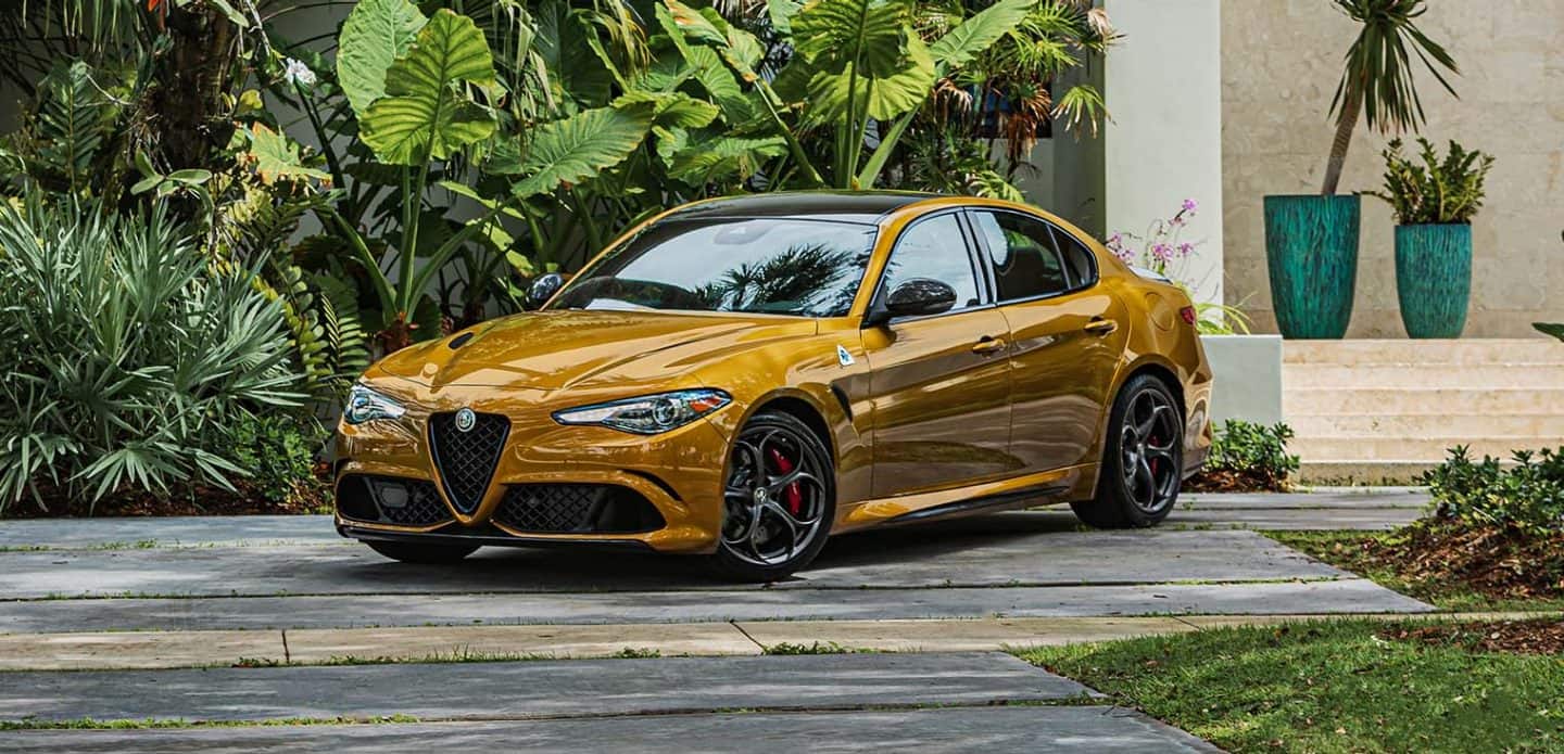 Display A three-quarter front view of a gold 2022 Alfa Romeo Giulia Quadrifoglio parked in the driveway of a home with a lush garden.
