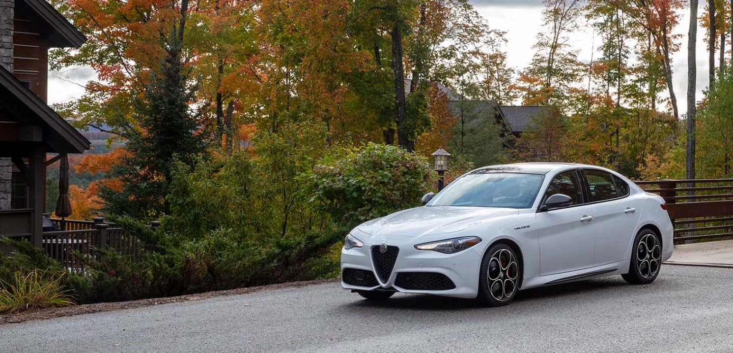 Display A white 2022 Alfa Romeo Giulia Veloce parked next to a cabin in the woods.