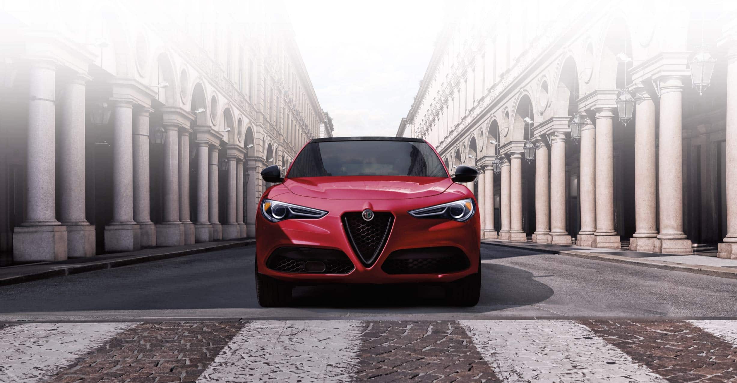 A front view of the 2022 Stelvio parked on a city street. Both sides of the street are lined with ornate columned building facades.