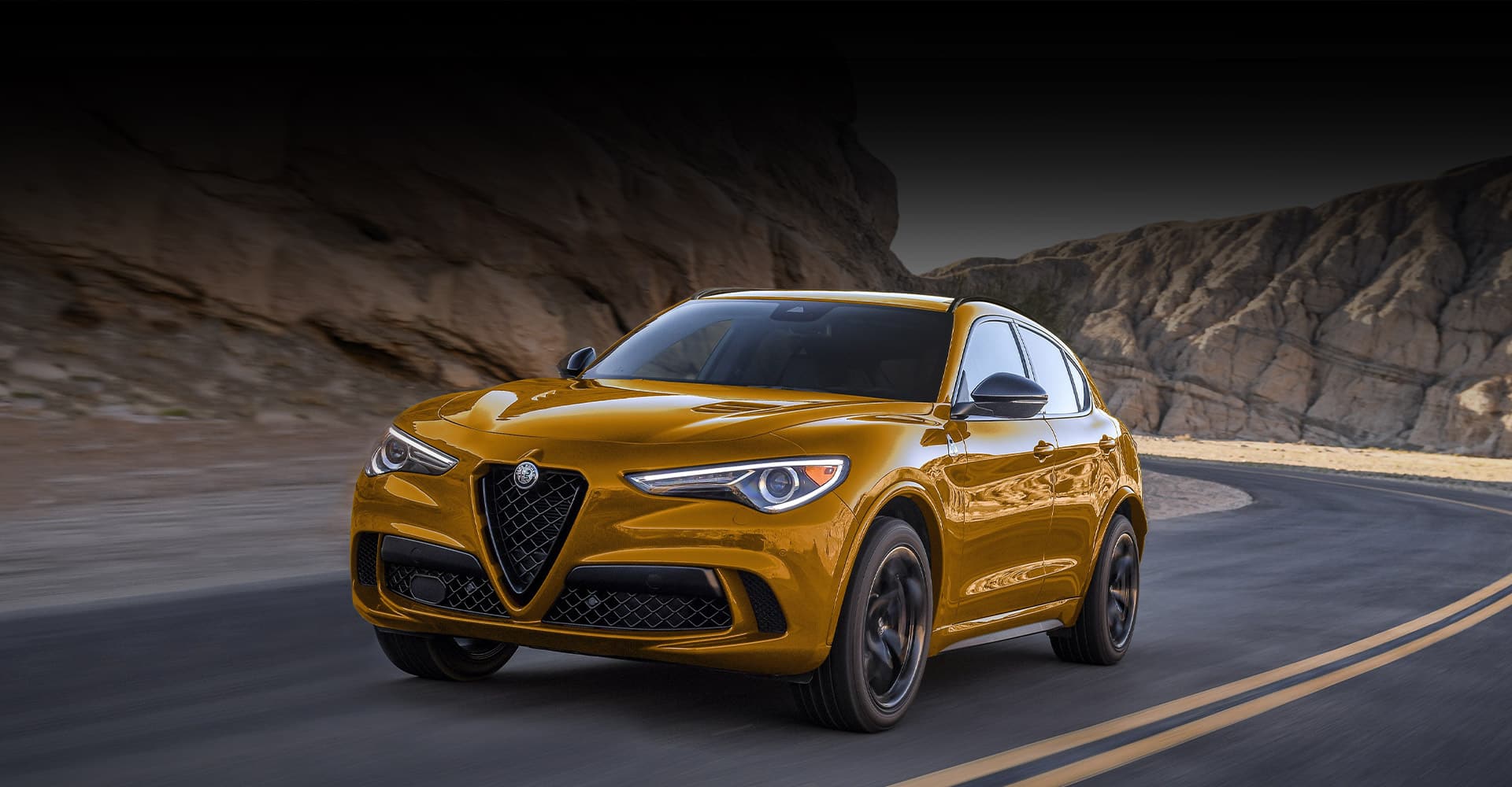 A gold 2022 Alfa Romeo Stelvio Quadrifoglio taking a curve at dusk, with jagged rock formations on both sides of the road.