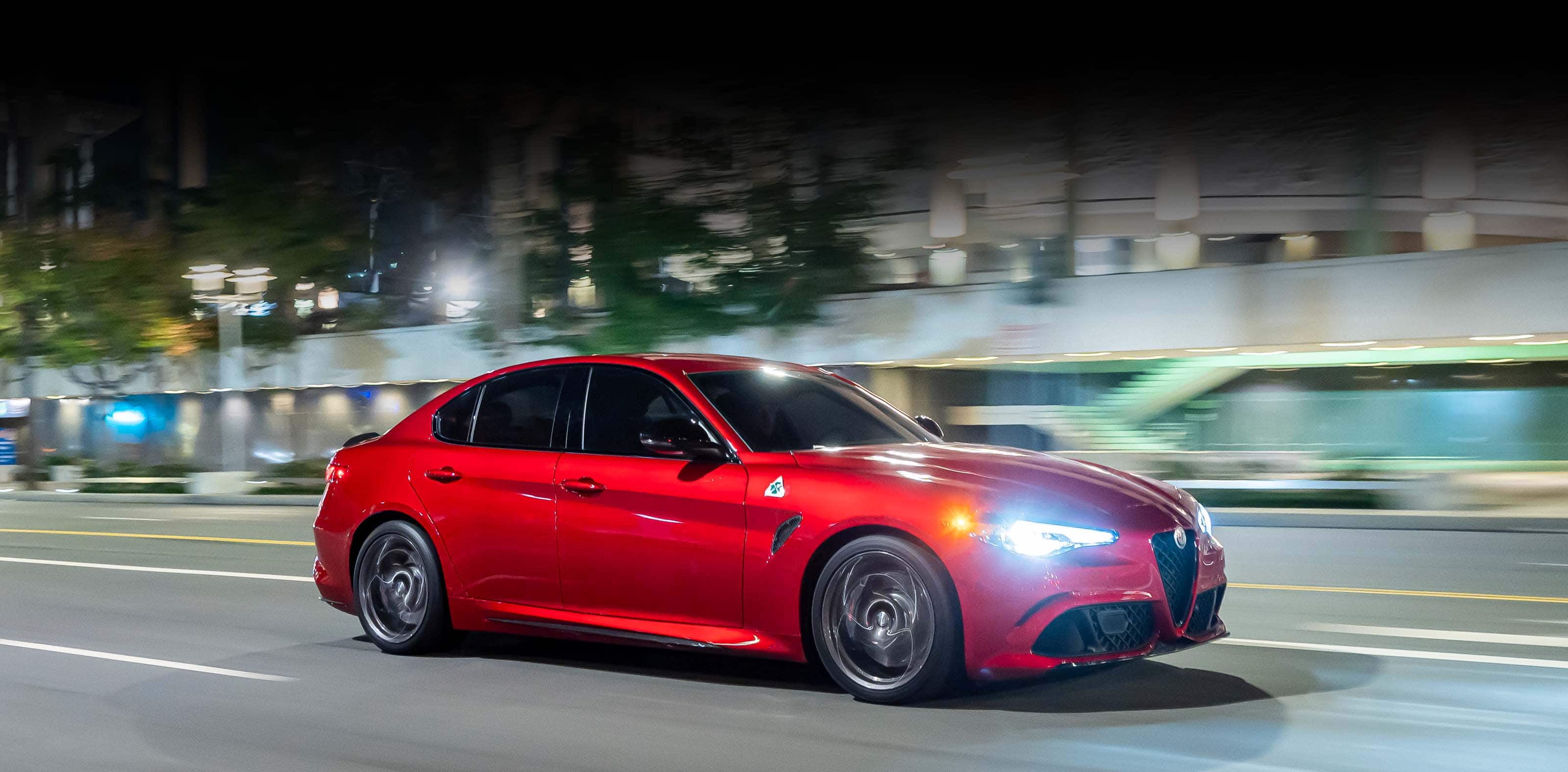 A passenger-side profile of a red 2023 Alfa Romeo Giulia Quadrifoglio being driven on a highway in the city at night. The background is blurred to indicate the speed of the vehicle.