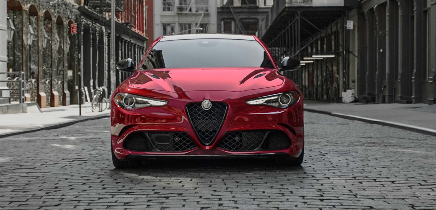 Display The front end on a red 2023 Alfa Romeo Giulia Quadrifoglio parked in the middle of a cobblestone city street with older buildings on three sides.