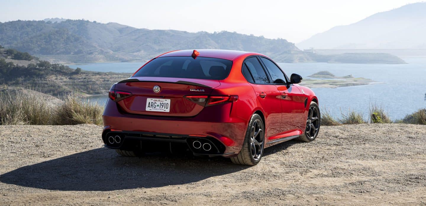 Display A rear view of a red 2023 Alfa Romeo Giulia Quadrifoglio parked on top of a drop-off, overlooking a body of water and a mountain in the distance.