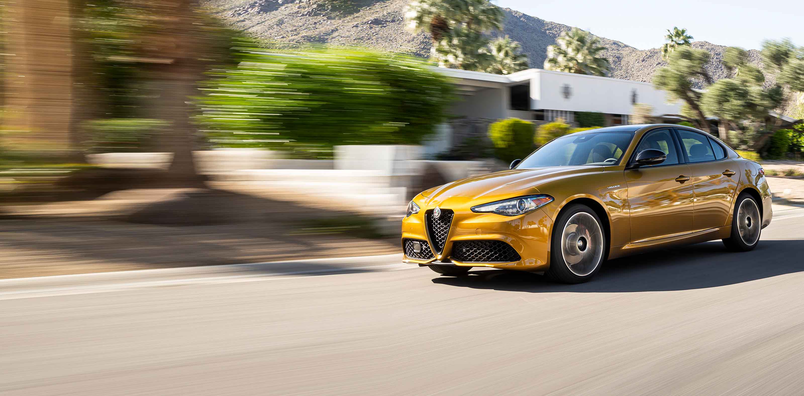 A gold 2023 Alfa Romeo Giulia Veloce being driven on a street with mountains in the distance, the background is blurred to indicate the speed of the vehicle.