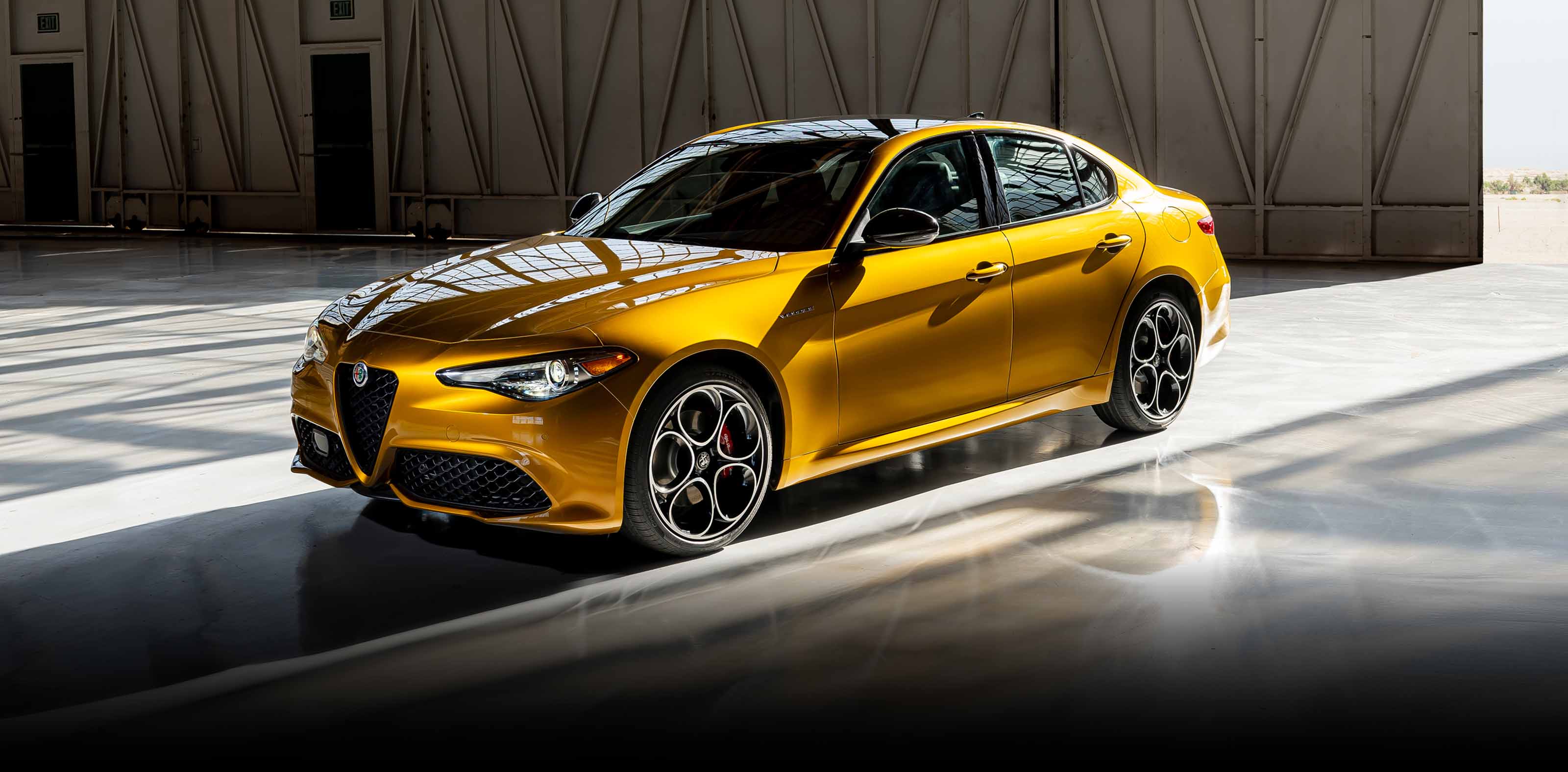 A gold 2023 Alfa Romeo Giulia Veloce parked in an industrial garage with light streaming in through the garage's open door.