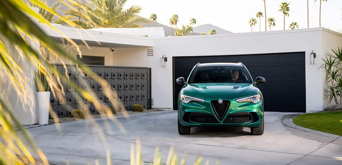Display The front view of a green 2023 Alfa Romeo Stelvio Veloce parked in the driveway of a contemporary home.