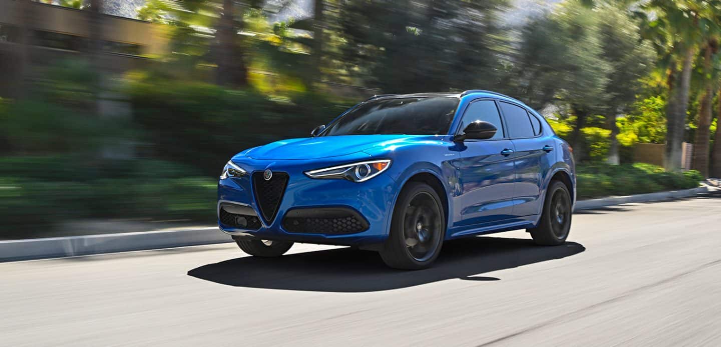 Display A blue 2023 Alfa Romeo Stelvio Veloce being driven on a road past a group of trees that are blurred, to demonstrate the speed of the vehicle.