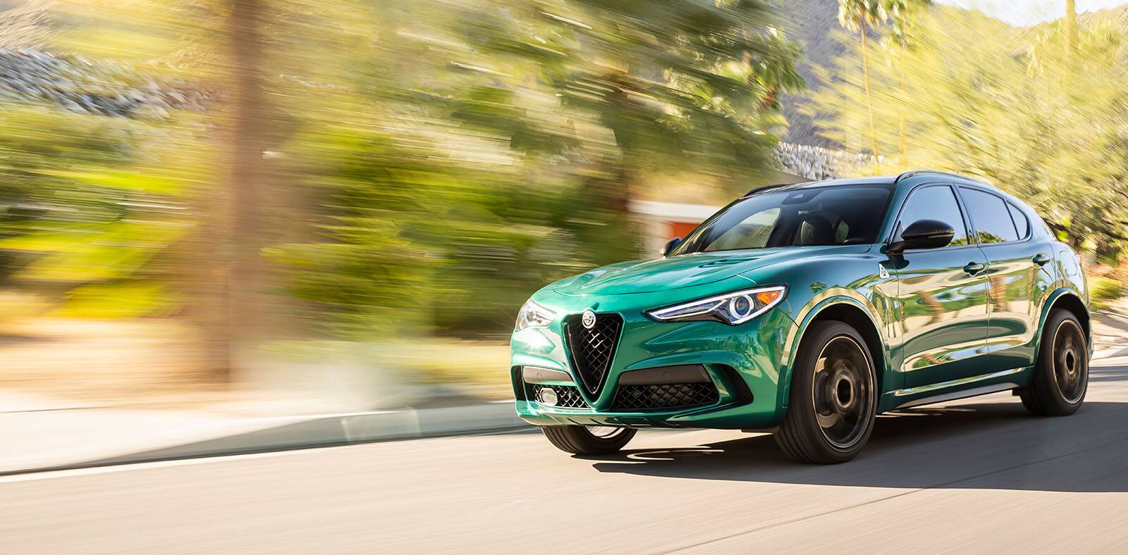 A green 2023 Alfa Romeo Stelvio Quadrifoglio being driven on a street with mountains in the distance, the background is blurred to indicate the speed of the vehicle.