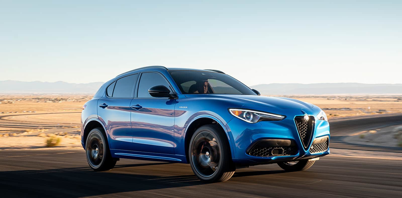 A blue 2023 Alfa Romeo Stelvio TBD being driven on a winding road in the desert.