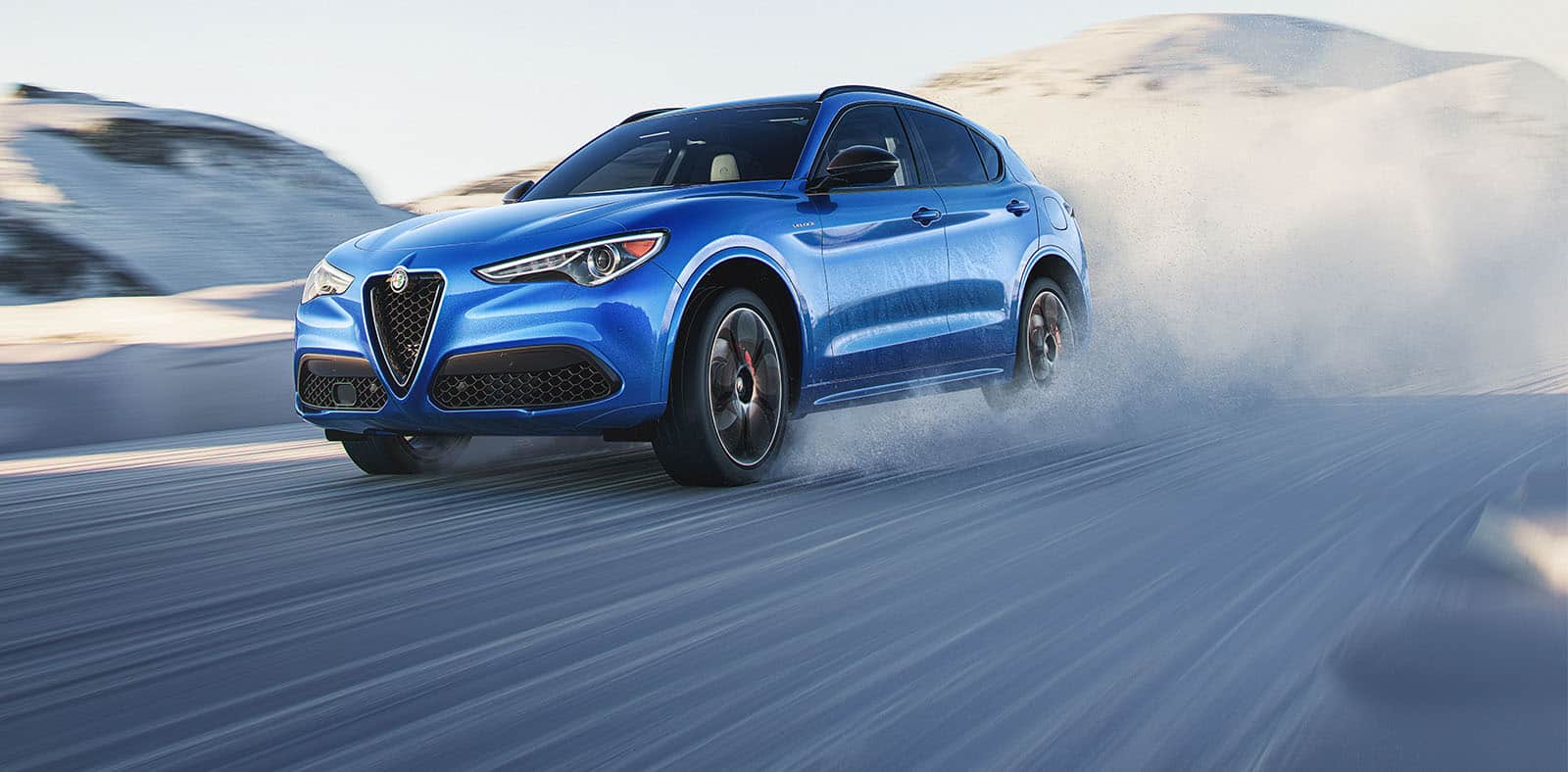 A blue 2023 Alfa Romeo Stelvio Veloce being driven on a snow-covered surface with the background blurred to indicate the speed of the vehicle.