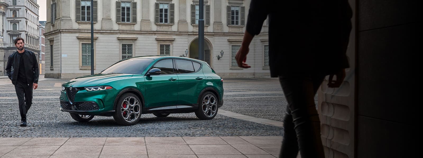 An angled driver-side profile of a green 2023 Alfa Romeo Tonale Veloce parked in a courtyard beside a classically-designed building with a woman walking toward the vehicle and a man walking beside it.