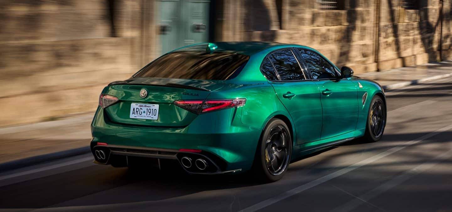 Display An angled rear passenger-side profile of a green 2024 Alfa Romeo Giulia Quadrifoglio being driven on a city street with the background blurred, indicating the vehicle is in motion.