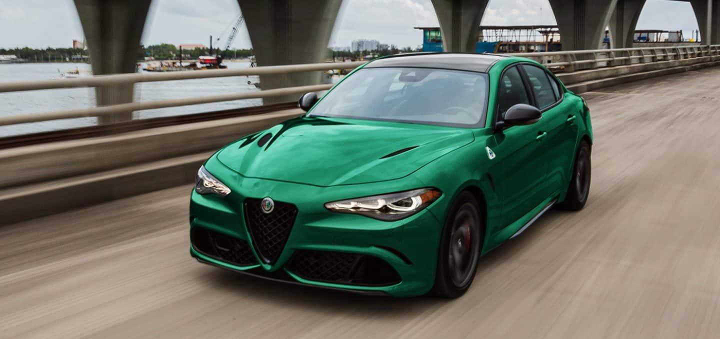 Display An angled front profile of a green 2024 Alfa Romeo Giulia Quadrifoglio being driven on a bridge over a body of water.