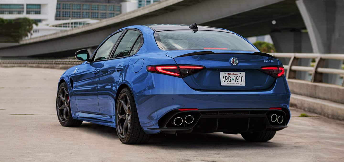 Display An angled rear driver-side profile of a blue 2024 Alfa Romeo Giulia being driven on a highway near an overpass.