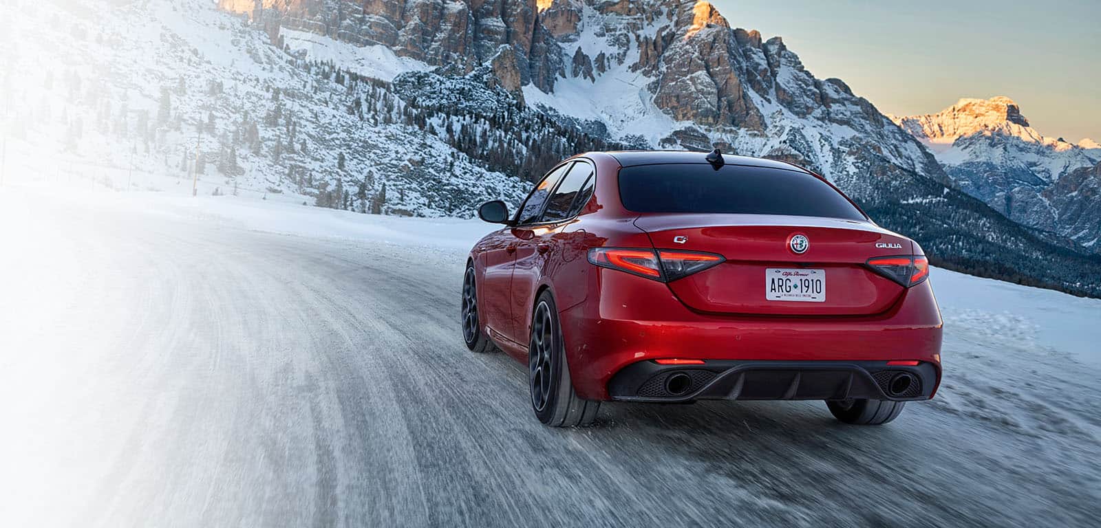 A rear angle of a red 2024 Alfa Romeo Giulia being driven up a snow-covered road in the mountains.