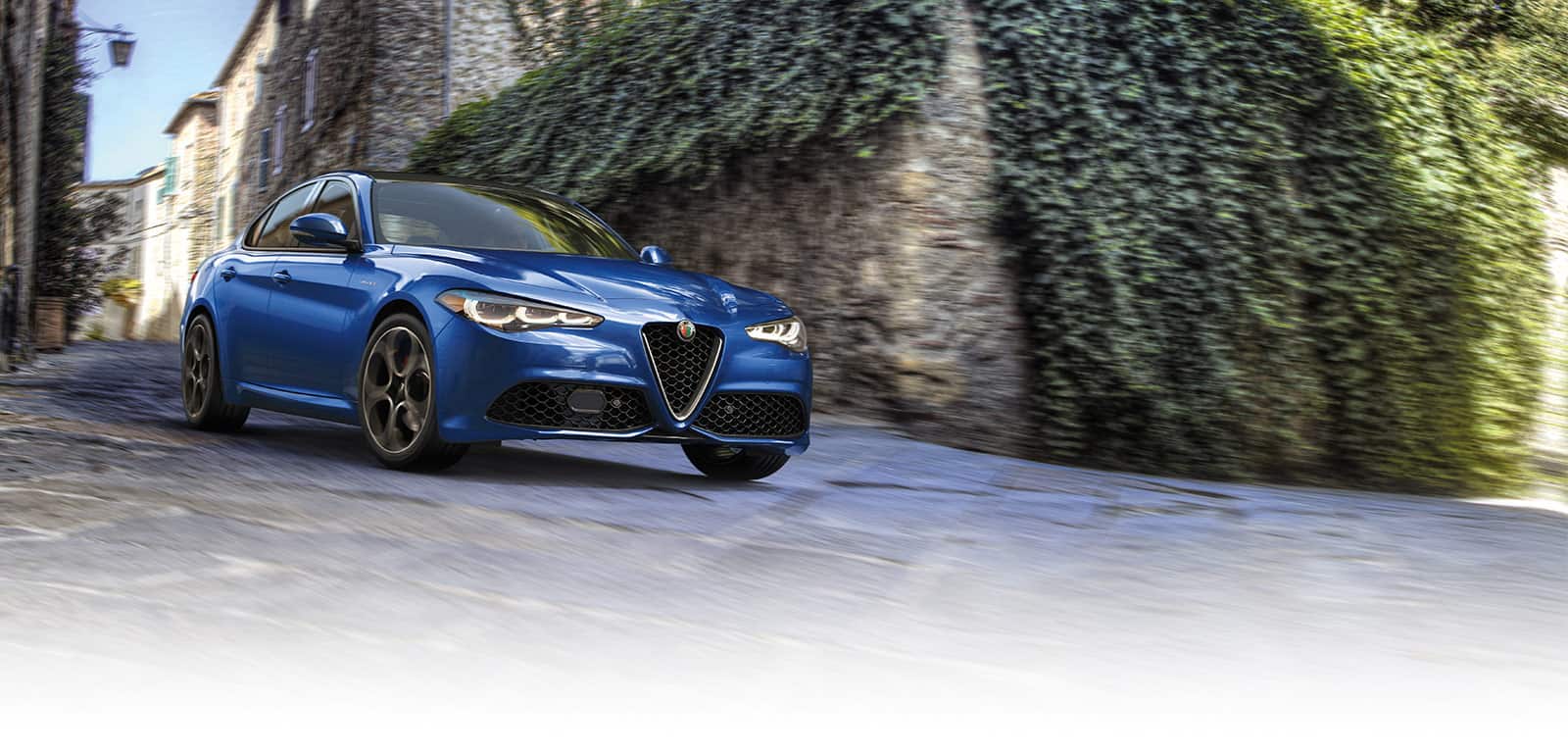 A blue 2024 Alfa Romeo Giulia being driven down a curved road in an area of classic European designed buildings. The background is blurred to indicate the vehicle is in motion.