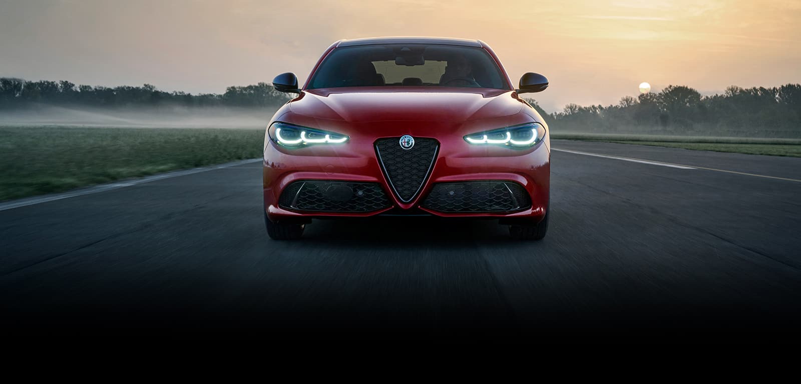 The front end of a red 2024 Alfa Romeo Giulia Veloce being driven on a track at sunset.