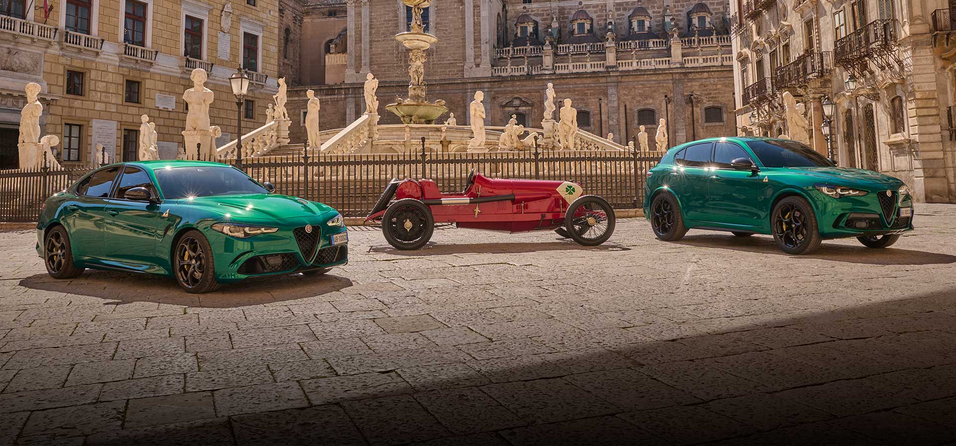 Three Alfa Romeo Quadrifoglio vehicles parked in a courtyard surrounded by Renaissance architecture. From left to right--a green 2024 Alfa Romeo Giulia Quadrifoglio, a vintage red Alfa Romeo Quadrifoglio race car and a green 2024 Alfa Romeo Stelvio Quadrifoglio.