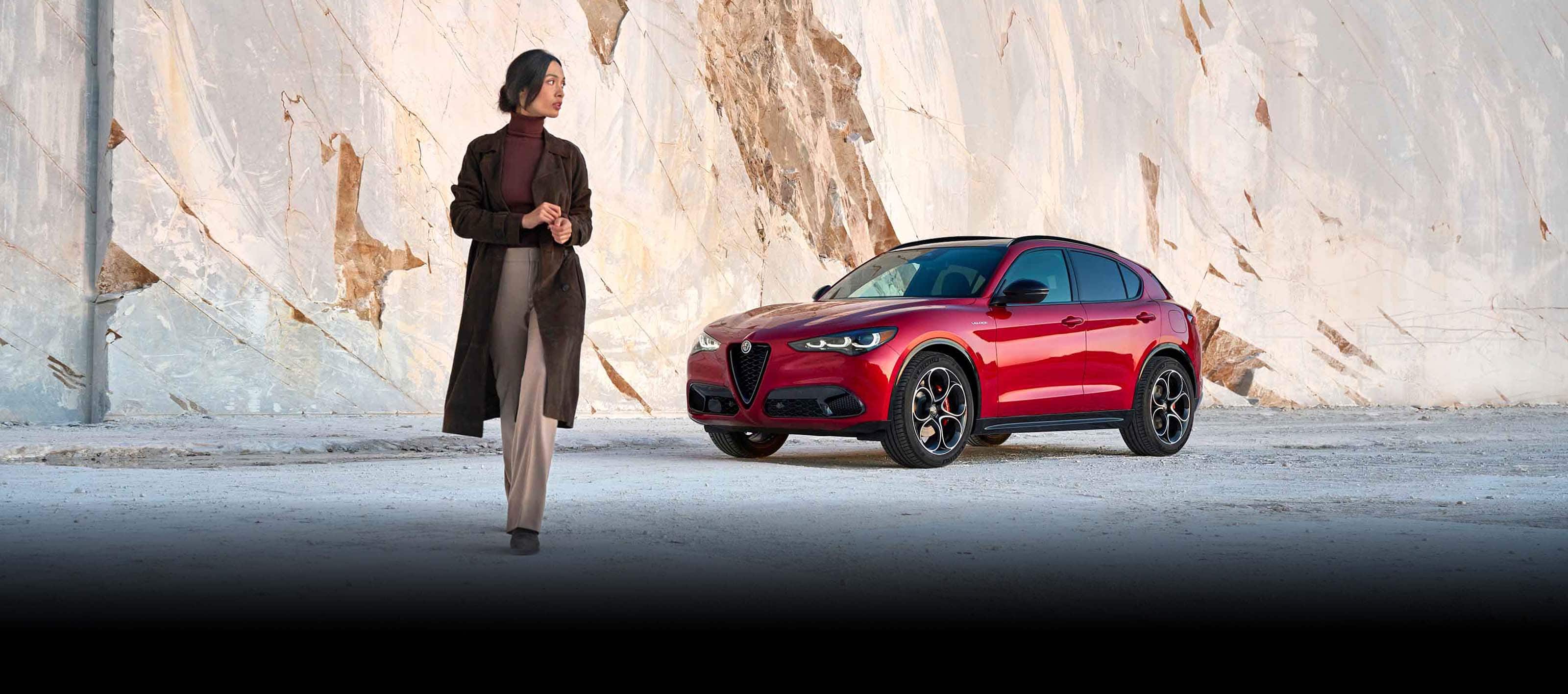 An angled front driver-side profile of red 2024 Alfa Romeo Stelvio TBD parked in front of a stone wall as a woman walks away from the vehicle.