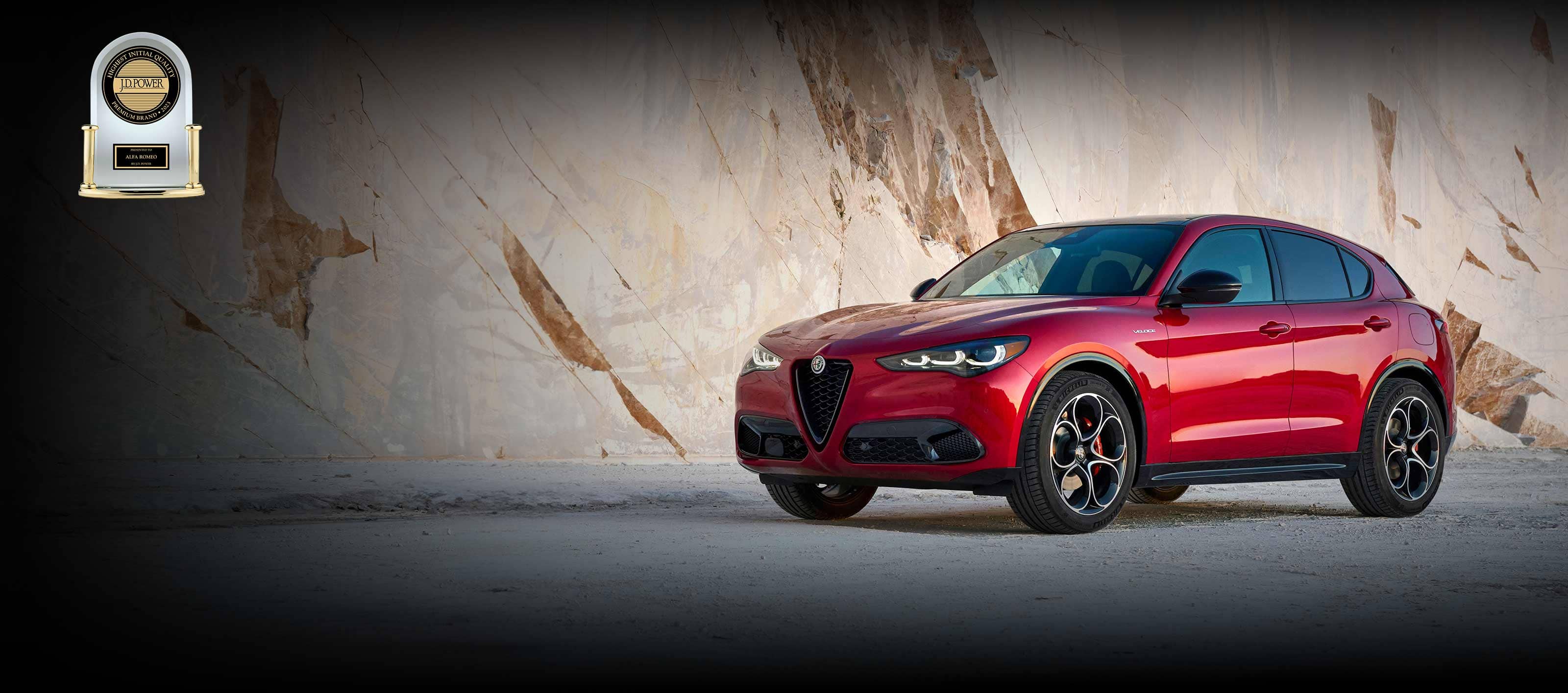 An angled driver-side profile of a red 2024 Alfa Romeo Stelvio Veloce displayed in front of a large marble wall. J.D. Power Highest Initial Quality Compact Premium SUV—2023 Presented to Alfa Romeo Stelvio by J.D. Power. J.D. Power Highest Initial Quality Premium Brand—2023 Presented to Alfa Romeo by J.D. Power.