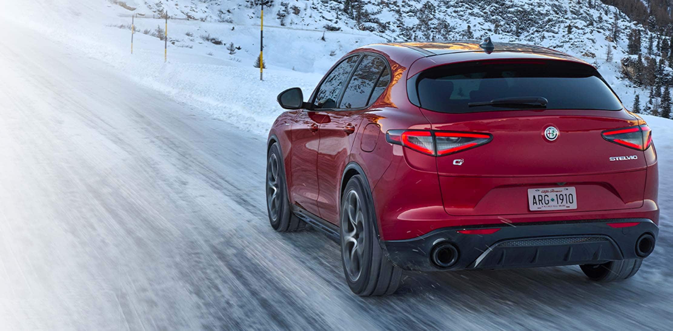 A rear angle of a red 2024 Alfa Romeo Stelvio TBD being driven up a snow-covered road in the mountains.