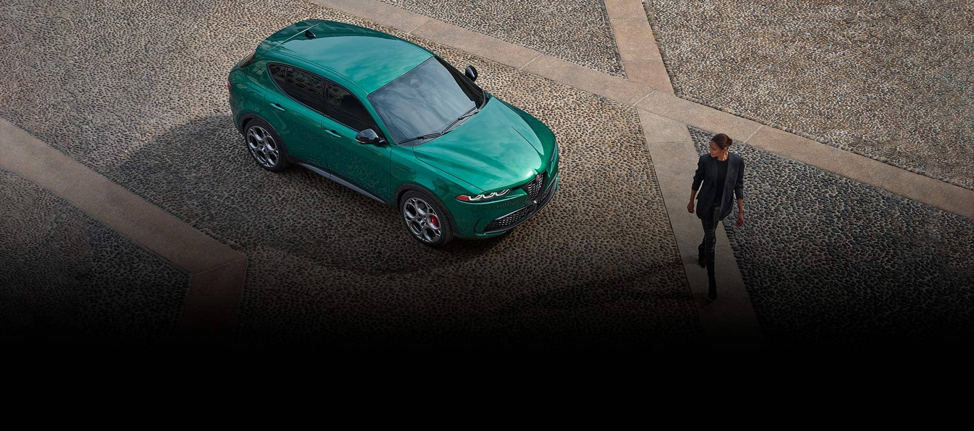An overhead angle of a green 2024 Alfa Romeo Tonale Veloce parked in a courtyard with a woman walking past as she looks at the vehicle.