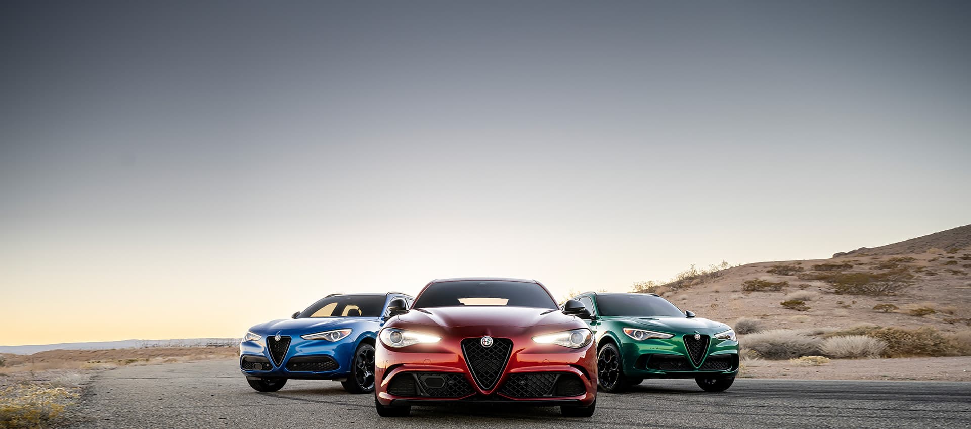 Three 2023 Alfa Romeo models parked beside a hill. From left to right: a blue 2023 Alfa Romeo Stelvio Quadrifoglio, a red 2023 Alfa Romeo Giulia Quadrifoglio and a green 2023 Alfa Romeo Stelvio Quadrifoglio.