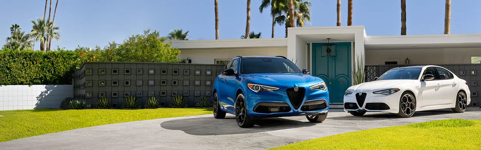 A blue 2022 Alfa Romeo Stelvio TBD and white 2022 Alfa Romeo Giulia TBD parked in the driveway of a contemporary home with palm trees in the background.