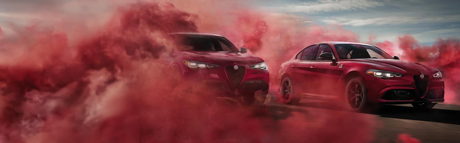 A red 2024 Alfa Romeo Stelvio Quadrifoglio and red 2024 Alfa Romeo Giulia Quadrifoglio side-by-side on a track, with red smoke enveloping both vehicles.