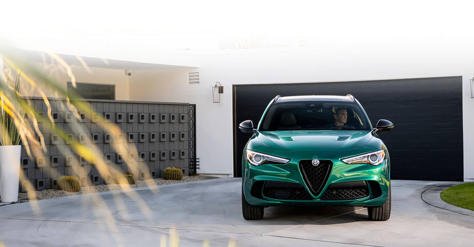 A green 2023 Alfa Romeo Stelvio Quadrifoglio parked in the driveway of a contemporary home with a driver inside.